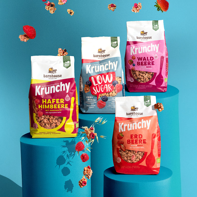 Blick von vorne auf die Beutel der vier Frucht-Krunchys Erdbeere, Hafer-Himbeere mit Amaranth, Low Sugar Very Berry sowie Waldbeere alternativ gesüßt von Bio-Knuspermüsli-Hersteller Barnhouse. Die Packungen stehen jeweils auf einem runden, blauen Sockel. Von oben fallen Knuspermüsli-Cluster, ein Haferhalm sowie Erdbeeren, Himbeeren und Heidelbeeren vor blauem Hintergrund nach unten.