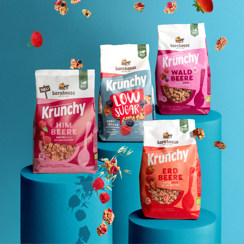 Blick von vorne auf die Beutel der vier Frucht-Krunchys Himbeere, Low Sugar Very Berry, Waldbeere alternativ gesüßt und Erdbeere von Bio-Knuspermüsli-Hersteller Barnhouse. Die Packungen stehen jeweils auf einem runden, blauen Sockel. Von oben fallen Knuspermüsli-Cluster, ein Haferhalm sowie Erdbeeren, Himbeeren und Heidelbeeren vor blauem Hintergrund nach unten.