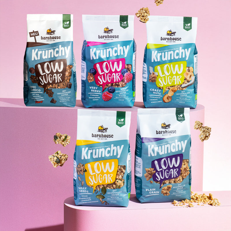 Blick von vorne auf die schräg angeordneten Packshots der fünf Low Sugar Krunchys Plain Grain, Crazy Nuts, Very Berry (No) Choco und Silly Seeds von Bio-Knuspermüsli-Hersteller Barnhouse, die auf zwei verschiedenen rosa Sockeln stehen. Außenrum fliegen Cluster.