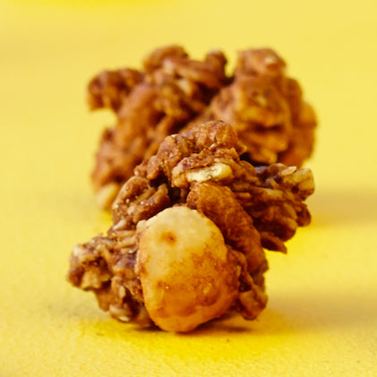 Großaufnahme von 2 Knuspermüsli-Clustern vor gelbem Hintergrund. Beim vorderen Cluster ist eine Haselnuss mit eingebacken.