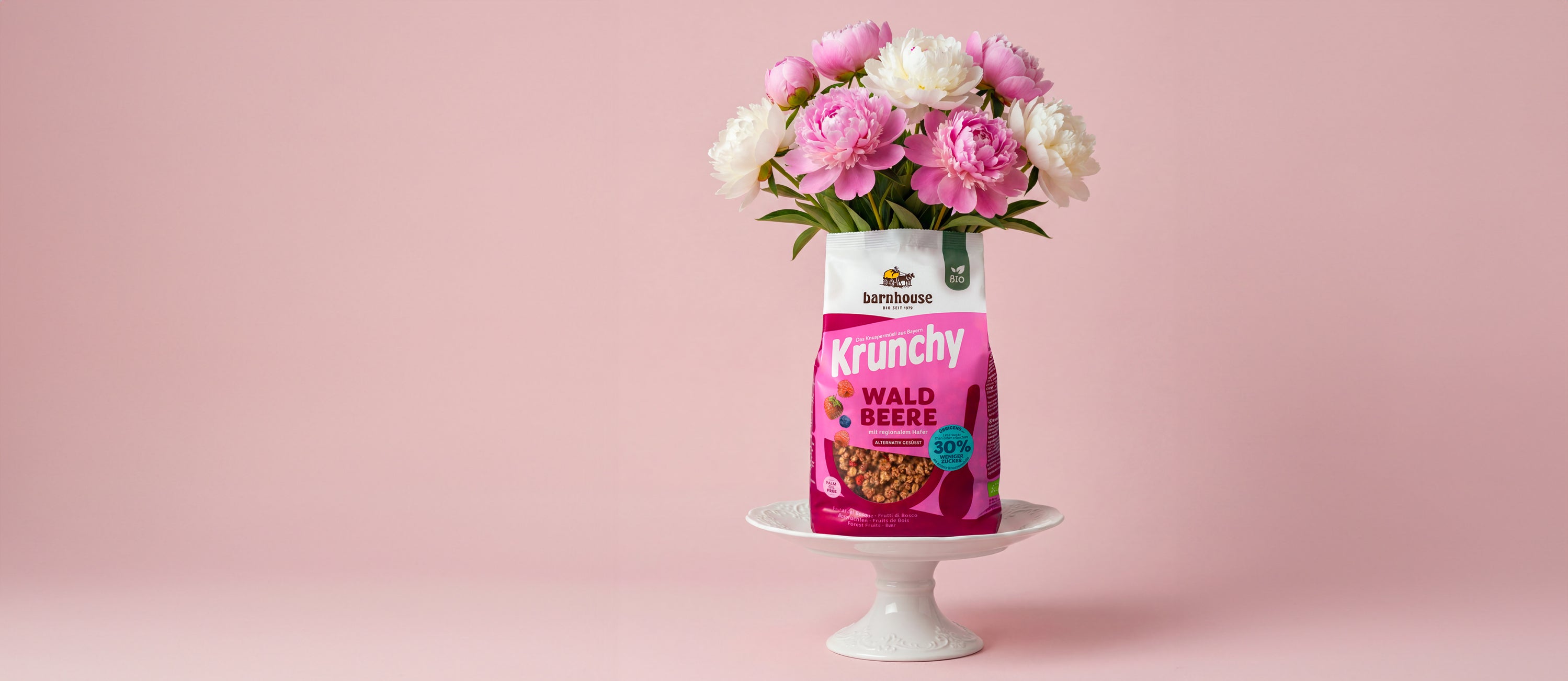 Beerenfarbener Beutel Krunchy Waldbeere weniger Zucker, aus dem ein Blumenstrauß mit pinken und weißen Blüten wächst. Das Bio-Knuspermüsli steht auf einer weißen Servierplatte aus Porzellan.