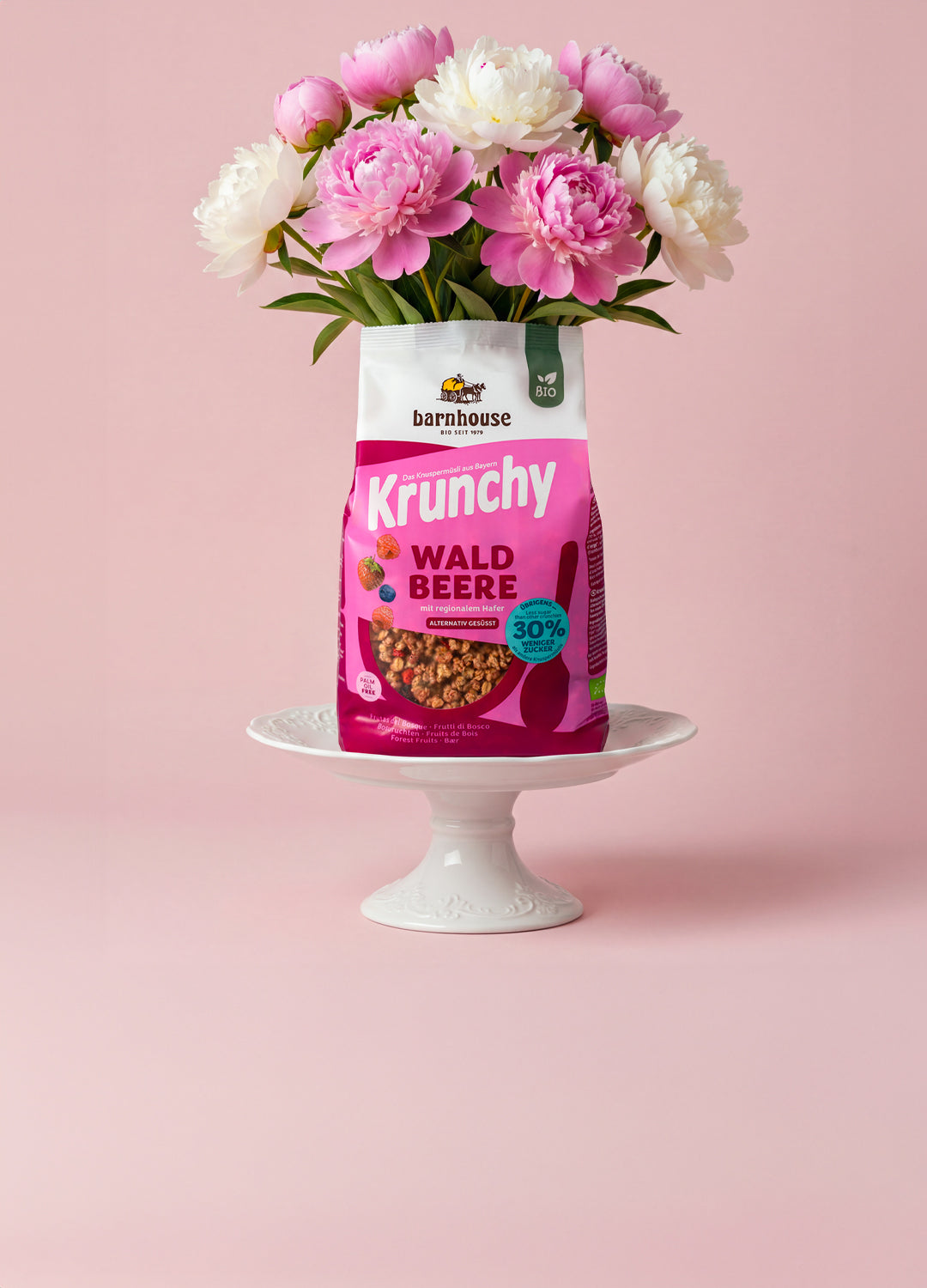Beerenfarbener Beutel Krunchy Waldbeere weniger Zucker, aus dem ein Blumenstrauß mit pinken und weißen Blüten wächst. Das Bio-Knuspermüsli steht auf einer weißen Servierplatte aus Porzellan.