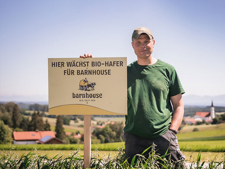 Barnhouse Bauer, der mit grünem T-Shirt und Arbeitshose in seinem Feld steht. Seine rechte Hand liegt auf dem Schild mit der Aufschrift "HIER WÄCHST BIO-HAFER FÜR BARNHOUSE". Im Hintergrund ist eine schöne Landschaft zu sehen.