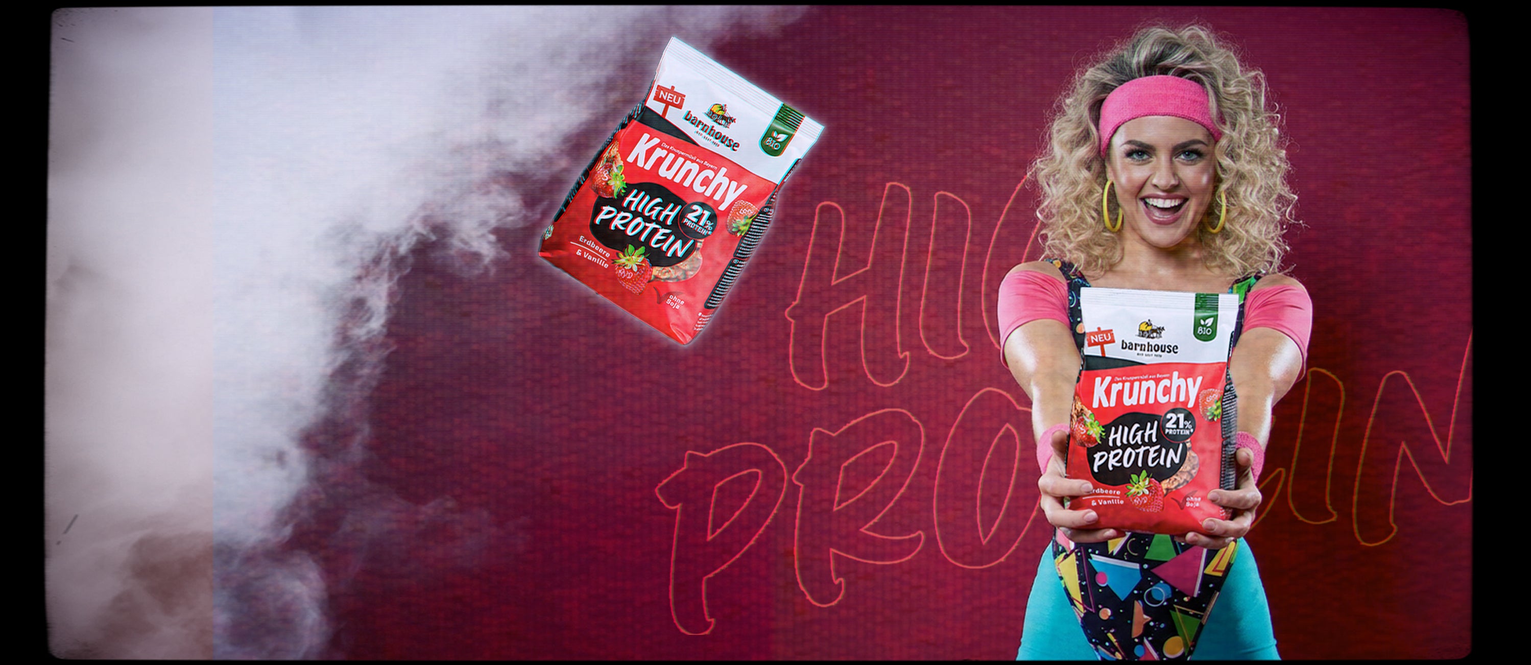 Frau mit bunten Sportklamotten aus den 80er Jahren, die freudestrahlend einen Beutel Krunchy High Protein in ihren Händen hält.