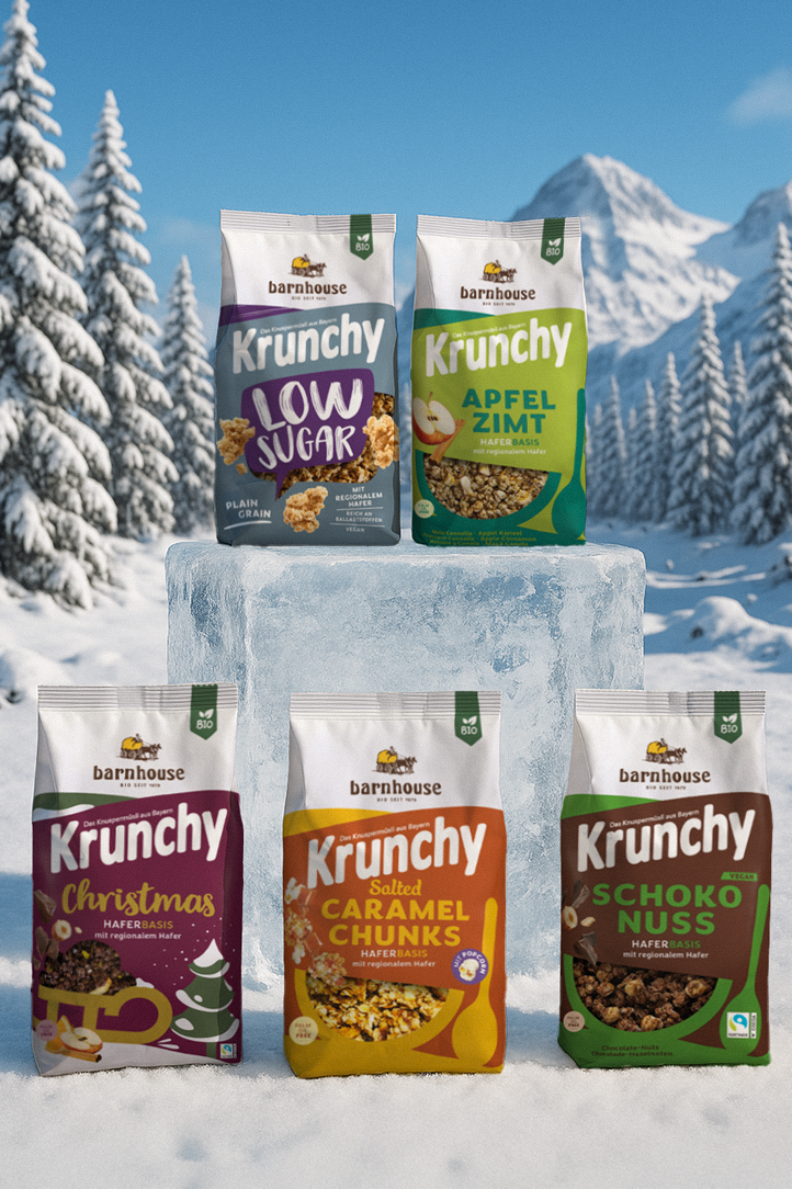 Winterlandschaft mit Bergen und Tannenbäumen, die mit Schnee bedeckt sind. In der Mitte steht ein Eisklotz, darauf die Beutel von Krunchy Low Sugar Plain Grain und Apfel-Zimt. Davor stehen die Beutel Krunchy Christmas, Salted Caramel Chunks und Schoko-Nuss.