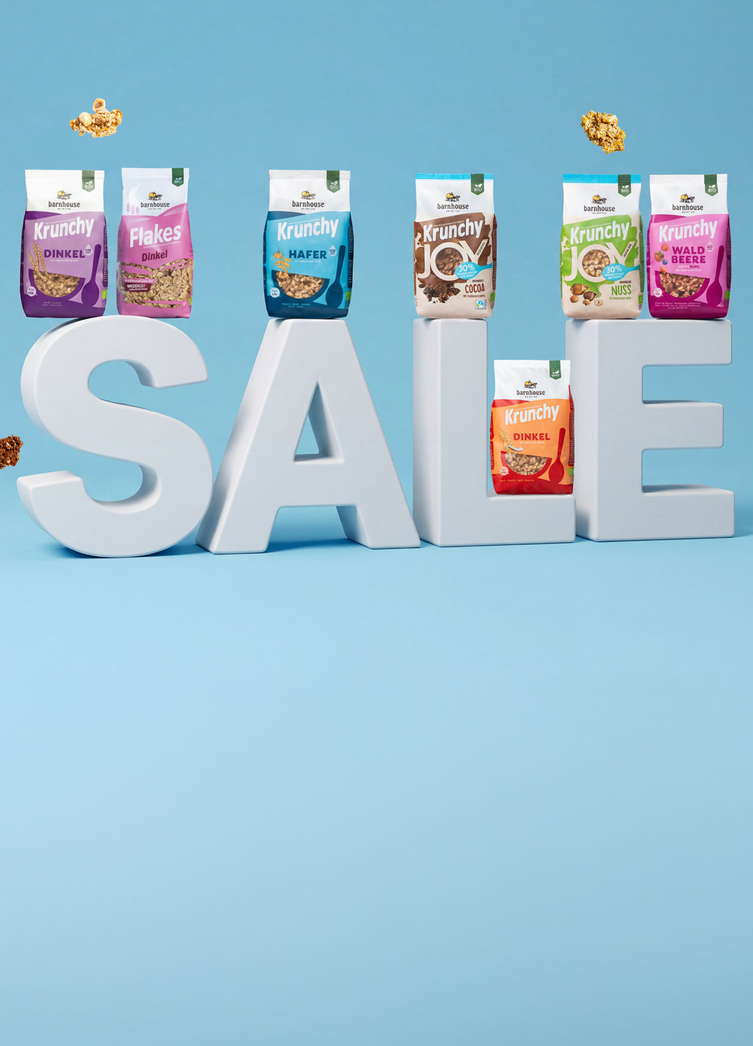SALE-Schriftzug, auf dem sieben verschiedene Krunchy-Packungen von Barnhouse stehen. Außenrum fliegen Bio-Knuspermüsli Cluster.