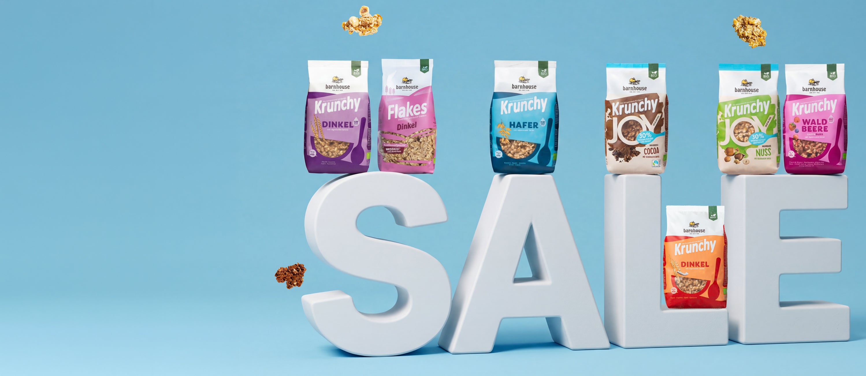 SALE-Schriftzug, auf dem sieben verschiedene Krunchy-Packungen von Barnhouse stehen. Außenrum fliegen Bio-Knuspermüsli Cluster.