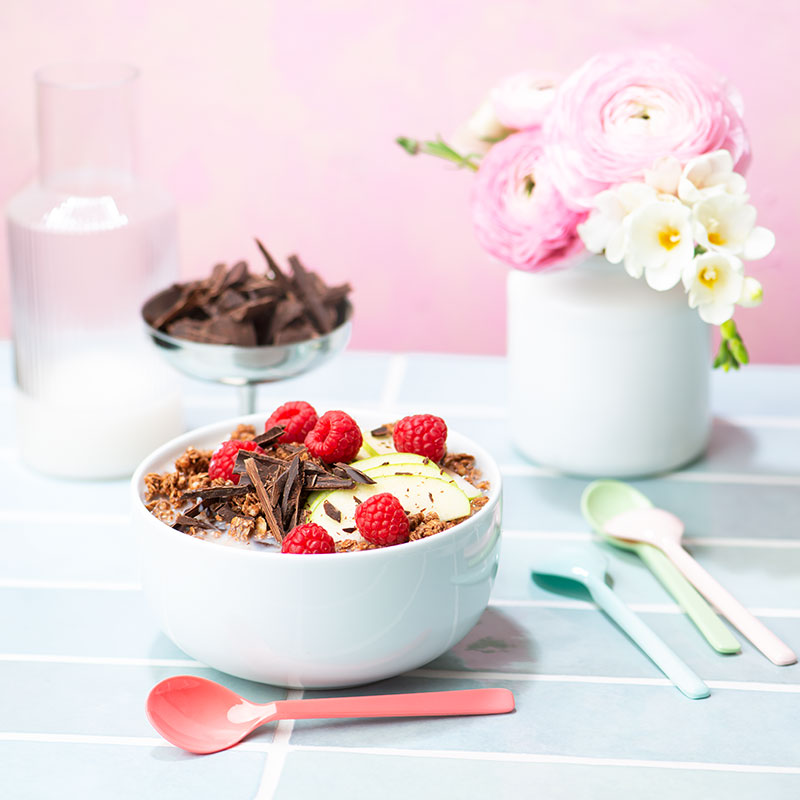Blick von vorne auf einen Frühstückstisch mit einer weißen Müslischale, die mit Milch und Krunchy Schoko von Bio-Knuspermüsli-Hersteller Barnhouse befüllt ist. Als dekoratives Highlight liegen noch Schokosplitter, Himbeeren und grüne Apfelscheiben mit drauf. Auf dem gefliesten Tisch steht eine silberne Schale mit Schokosplittern sowie eine weiße Vase mit rosa und weißen Blumen. Vorne liegen noch vier Löffel in verschiedenen Farben.