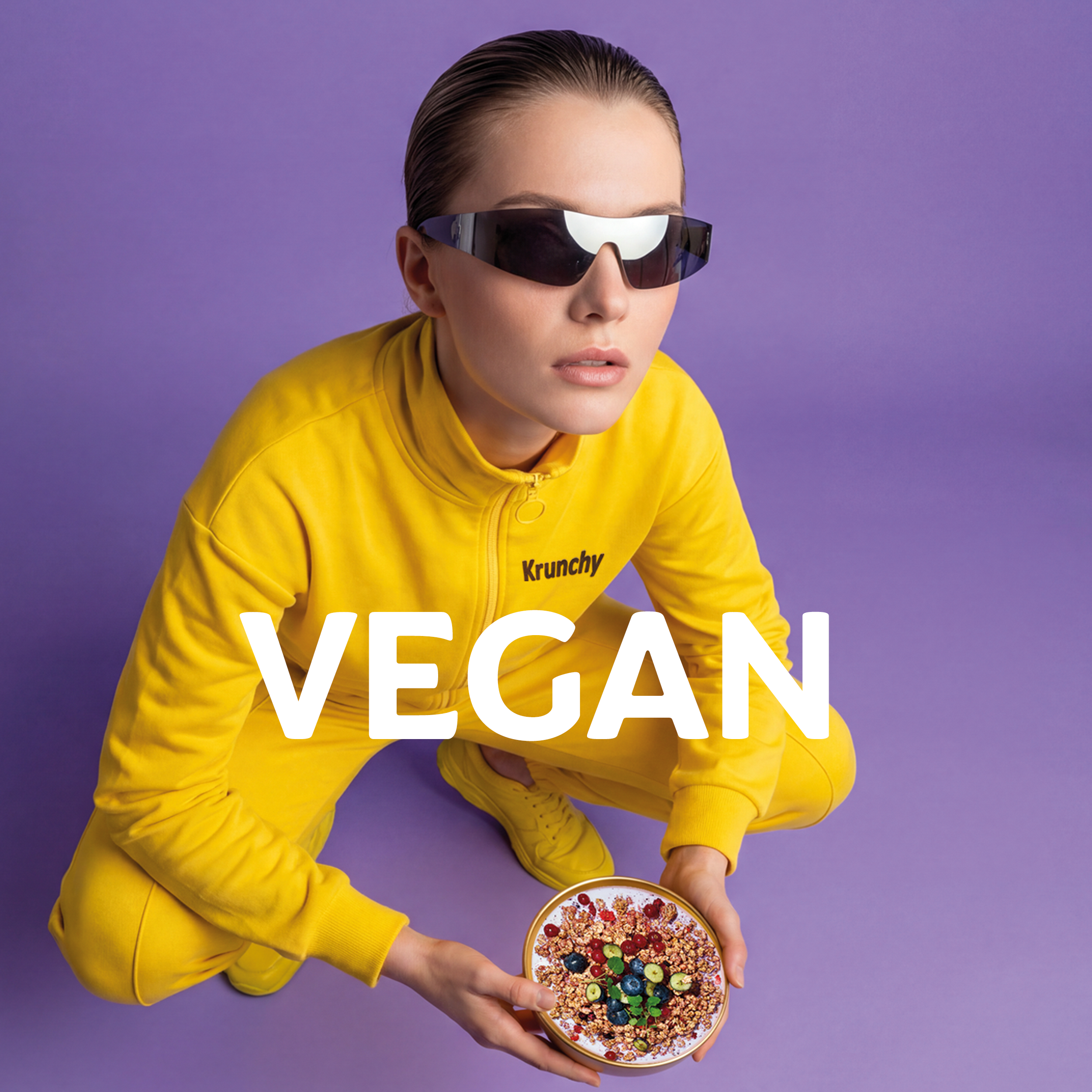 Dunkelhaarige Frau mit Sonnenbrille und gelbem Krunchy-Trainingsanzug, die vor einem lilafarbenen Hintergrund in der Hocke kniet. Sie schaut ernst in die Kamera und hält mit ihren Händen eine goldene Schale mit Bio-Knuspermüsli, Milch und verschiedenen Beeren. In der Mitte des Bilder ist der Schriftzug "VEGAN" zu sehen.