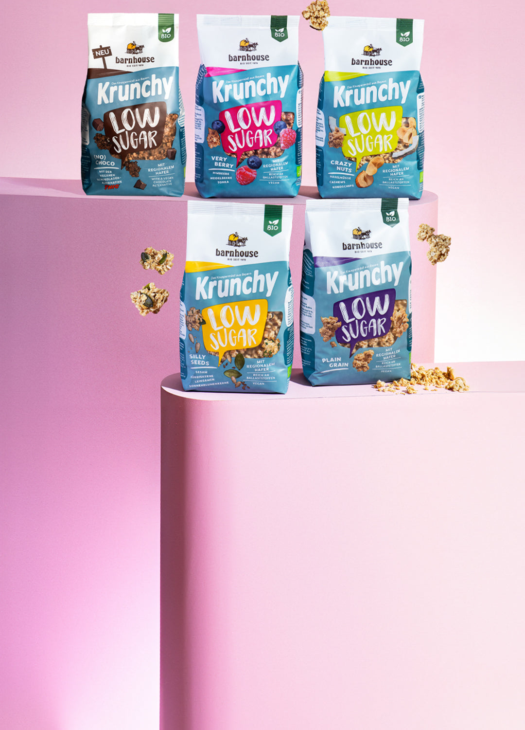 Blick von vorne auf die fünf Low Sugar Krunchys Plain Grain, Crazy Nuts, Very Berry (No) Choco und Silly Seeds, die auf zwei verschiedenen rosa Sockeln stehen. Außenrum fliegen Cluster.