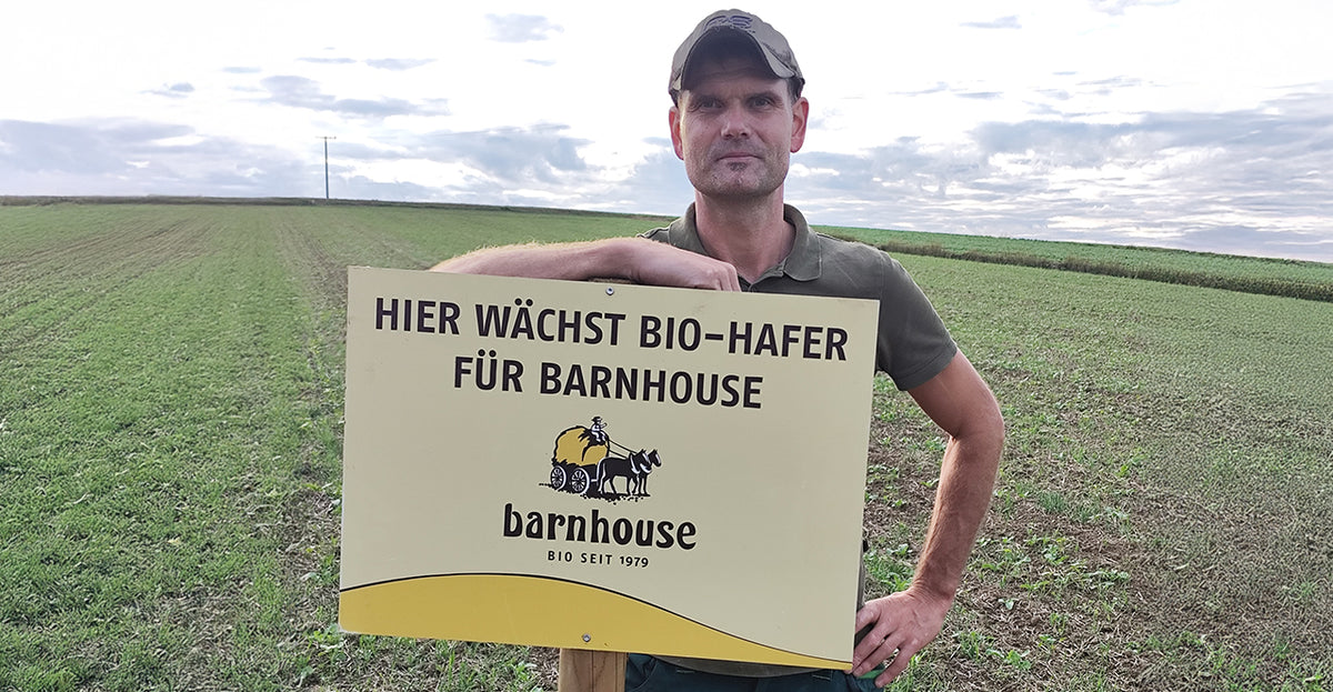 Barnhouse Bauer Tobias Oberwallner der Lebensgemeinschaft Höhenberg steht in seinem Feld, auf dem bald Hafer für Bio-Knuspermüsli von Barnhouse wächst. Er lehnt an einem Schild mit Barnhouse-Logo und der Aufschrift "Hier wächst Bio-Hafer für Barnhouse".