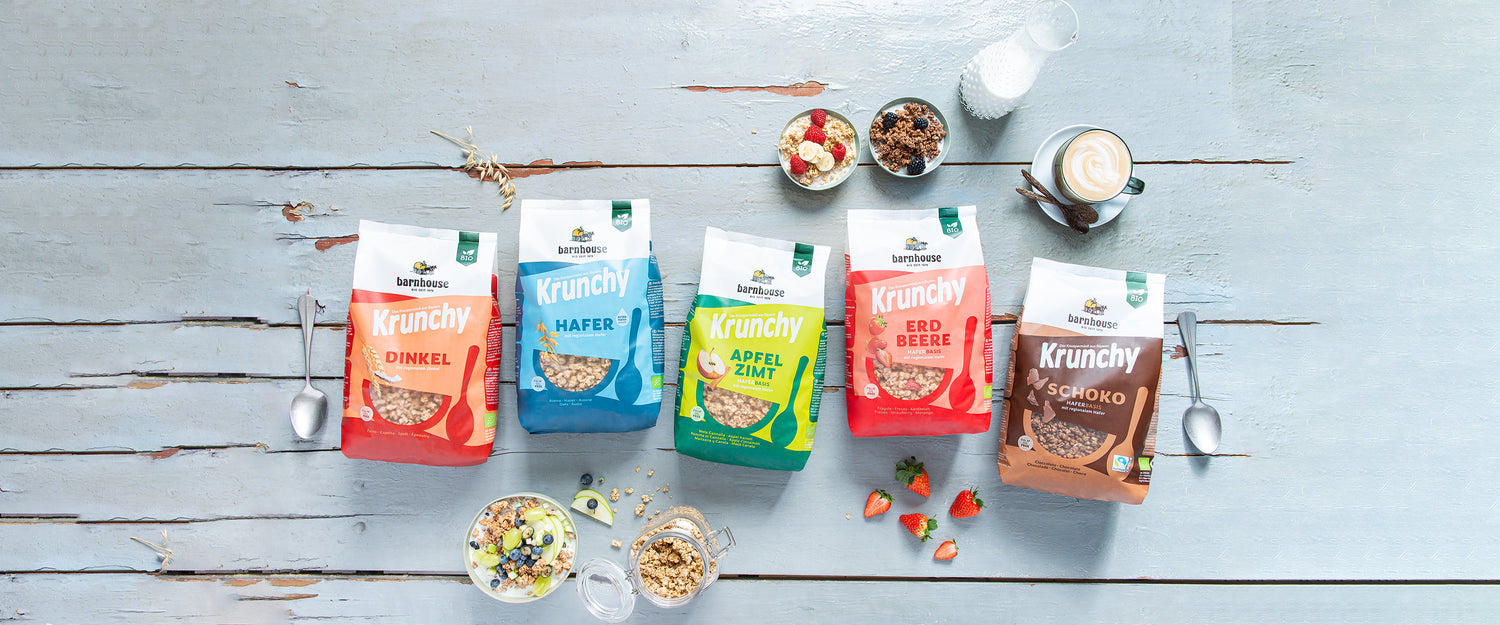 Blick von oben auf fünf verschiedene Krunchy-Beutel von Bio-Knuspermüsli-Hersteller Barnhouse, die mit verschiedenen Müslischalen, Früchten, Cappuccino und Milch auf einem blauen gestrichenen Holztisch liegen.