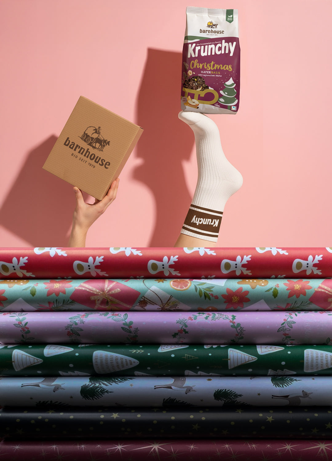 Hand vor einem rosa Hintergrund, die einen braunen Karton mit Barnhouse Logo hält. Daneben ein Fuß mit weiß-brauner Barnhouse-Socke, dir einen Beutel Krunchy Christmas balanciert. Unten buntes Geschenkpapier.