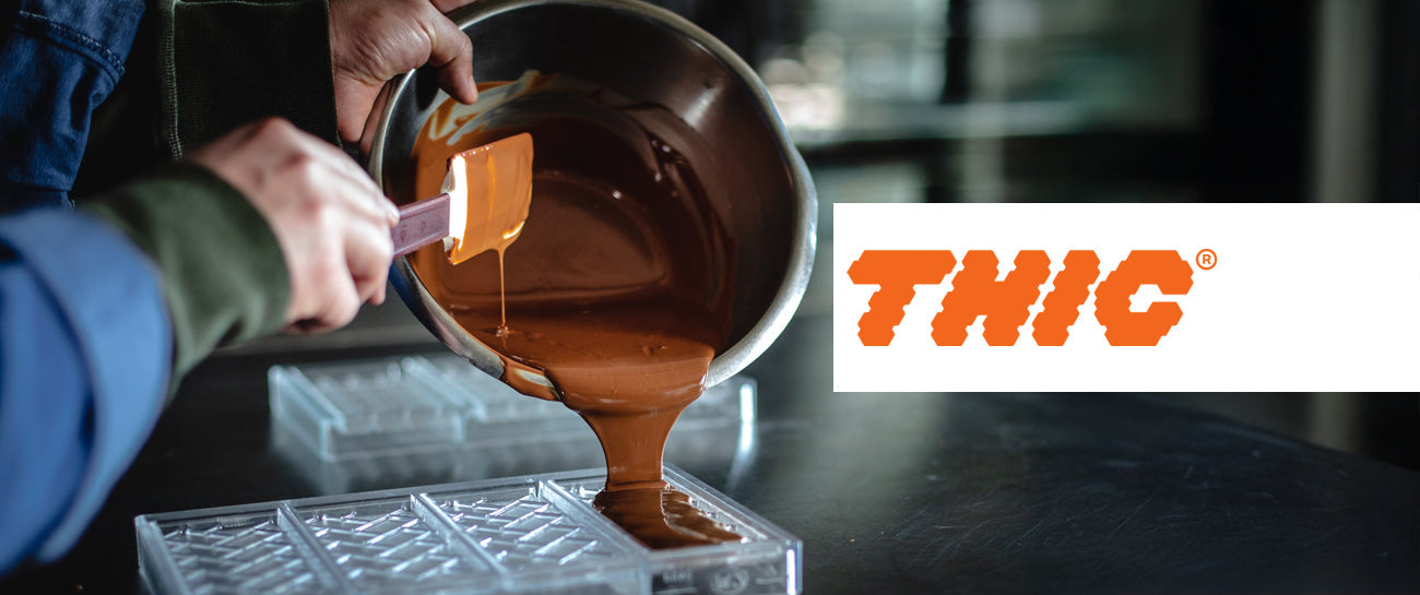 Eine Person gießt geschmolzene Schokolade aus einer Metallschüssel in eine Schokoladenform. Rechts im Bild ist das orangefarbene Logo mit dem Schriftzug "THIC" auf weißem Hintergrund zu sehen.
