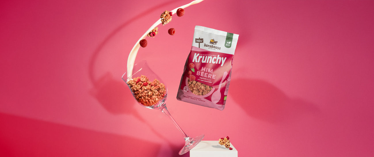 Beutel Krunchy Himbeere neben Weinglas, das mit Clustern des Himbeer-Müslis befüllt ist und vom weißen Sockel fällt. Von oben fließt ein Milchstrahl ins Glas, weitere Cluster & Himbeeren fliegen herum