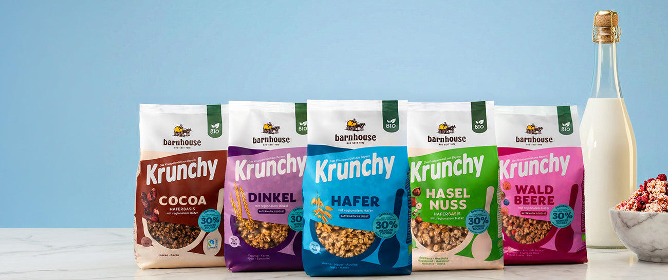5 Krunchys mit 30% weniger Zucker vor hellblauer Wand auf Marmorplatte: Cocoa, Dinkel, Hafer, Haselnuss und Waldbeere. Rechts daneben stehen eine Glasflasche mit Milch sowie eine Müslischüssel aus Marmor mit Krunchy Waldbeere.