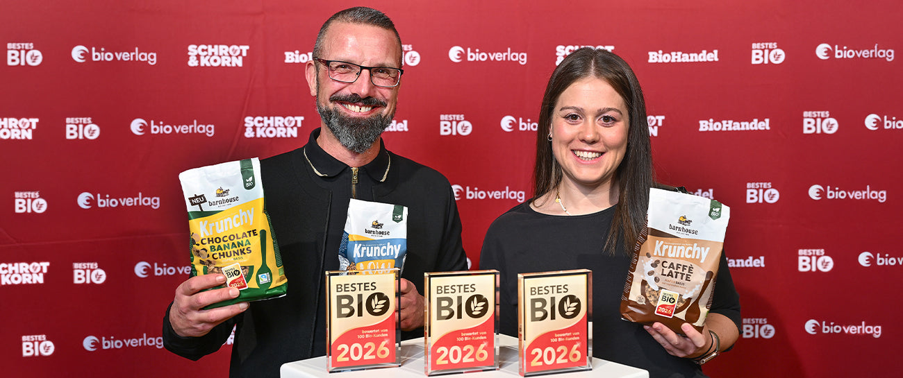 Andreas Bentlage und Kathi Schmidingerhalten die Beutel von Krunchy Chocolate Banana Chunks, Low Sugar Silly Seeds und Caffè Latte in die Kamera. Vor ihnen stehen die Trophäen der drei Krunchys für "Bestes Bio 2026".