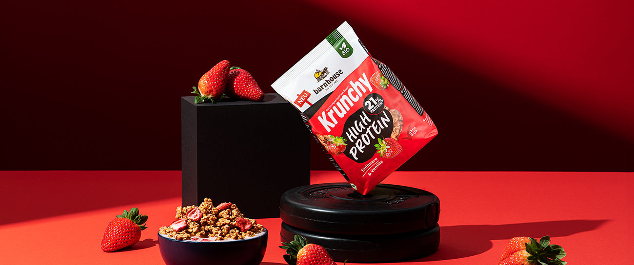 Beutel Krunchy High Protein von Barnhouse auf 2 Hantelgewichten. Links daneben ein schwarzes Achteck mit Erdbeeren. Vorne steht eine dunkle Schale mit dem Krunchy Erdbeere und Milch.