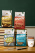 Holztisch, der vor einer grünen Tafel im Klassenzimmer steht. Auf dem Tisch liegen 2 Bleistifte, 1 Kugelschreiber und 1 Lineal. Dahinter stehen je eine XL-Packung Krunchy Low Sugar Crazy Nuts und Hafer alternativ gesüßt. Dahinter sind 2 Türme mit gestapelten Büchern, auf denen eine XL-Packung Krunchy Chocolate Chunks und Erdbeere stehen. Auf der rechten Seite steht ein befüllter Krunchy2go-Becher.