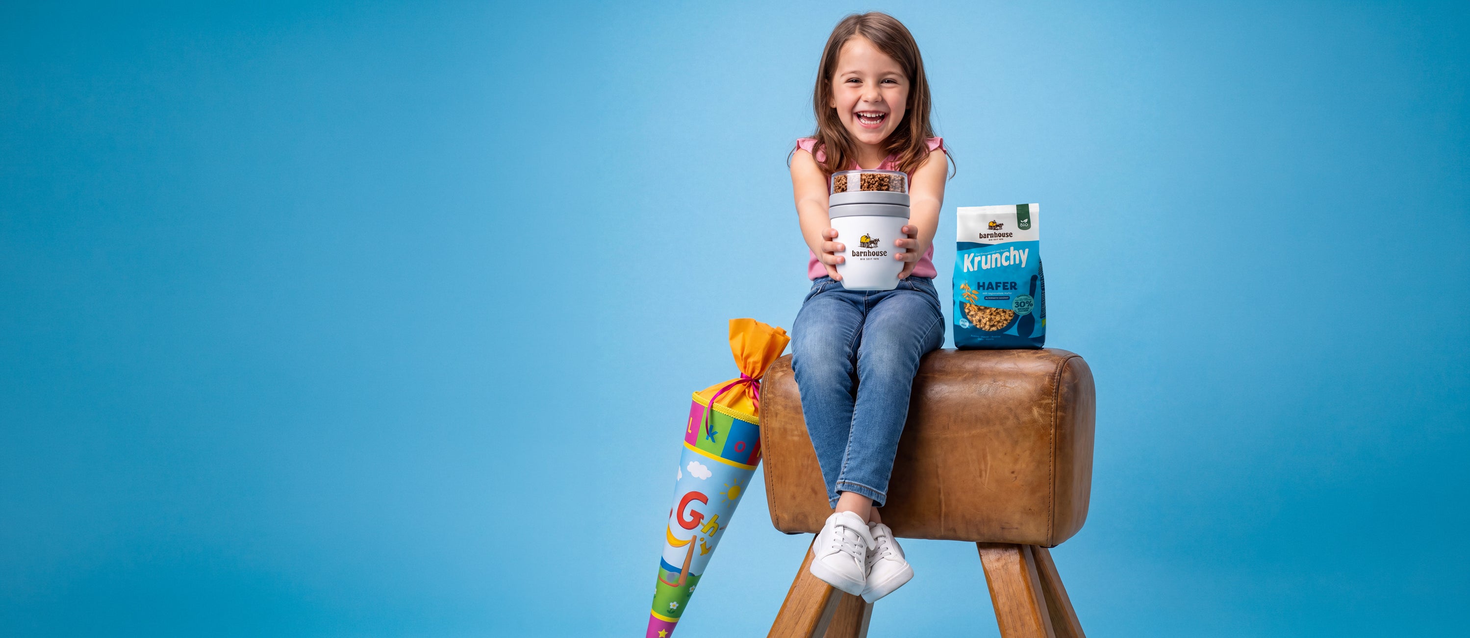 Braunhaariges Schulmädchen mit rosa Top, blauer Jeans und weißen Sneaker, das mit überschlagenen Beinen auf einem Sprungbock sitzt. Sie hält mit zwei ausgestreckten Armen einen Krunchy2go-Becher in die Kamera und grinst glücklich über beide Ohren. Neben ihr auf dem Bock steht ein blauer Beutel Krunchy Hafer alternativ gesüßt. Außerdem lehnt an dem Bock eine bunte Schultüte.