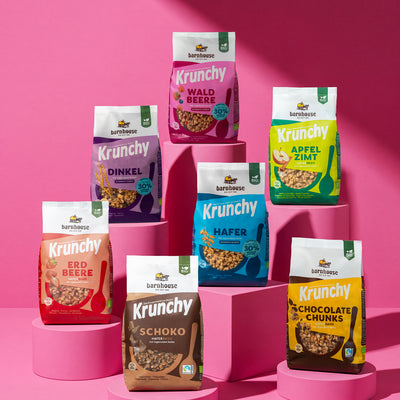 Bild von vorne auf alle Krunchys, die im Abo-Paket Family enthalten sind: Krunchy Schoko, Krunchy Hafer weniger Zucker,  Krunchy Dinkel weniger Zucker, Krunchy Erdbeere, Krunchy Waldbeere weniger Zucker, Krunchy Apfel-Zimt und Krunchy Chocolate Chunks. Hafer steht auf einem rosafarbenen, eckigen Sockel und alle weiteren Sorten auf rosafarbenen, runden Sockeln.