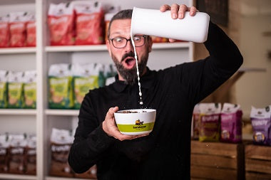 Spaßfoto: Andreas - Leitung Marketing & Produktmanagement - hält mit der rechten Hand eine grün-weiße Barnhouse-Schüssel und mit der linken Hand schüttet er aus großer Entfernung Milch in die Schale. Dabei hat er Munf und Augen weit aufgerissen. Hinter ihm steht ein Regal mit Krunchy von Bio-Knuspermüsli-Hersteller Barnhouse.