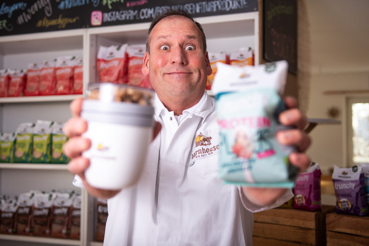 Spaßfoto: Axel - Leitung Produktion - reißt seine Augen auf und streckt grinsend einen Krunchys2go-Becher sowie einen Krunchy-Beutel in die Kamera. Hinter ihm steht ein Regal mit Krunchy von Bio-Knuspermüsli-Hersteller Barnhouse.
