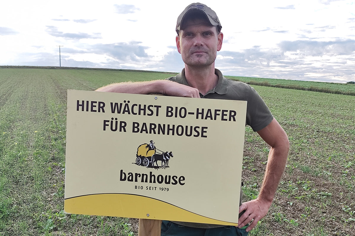 Barnhouse Bauer Tobias Oberwallner der Lebensgemeinschaft Höhenberg steht in seinem Feld, auf dem bald Hafer für Bio-Knuspermüsli von Barnhouse wächst. Er lehnt an einem Schild mit Barnhouse-Logo und der Aufschrift "Hier wächst Bio-Hafer für Barnhouse".