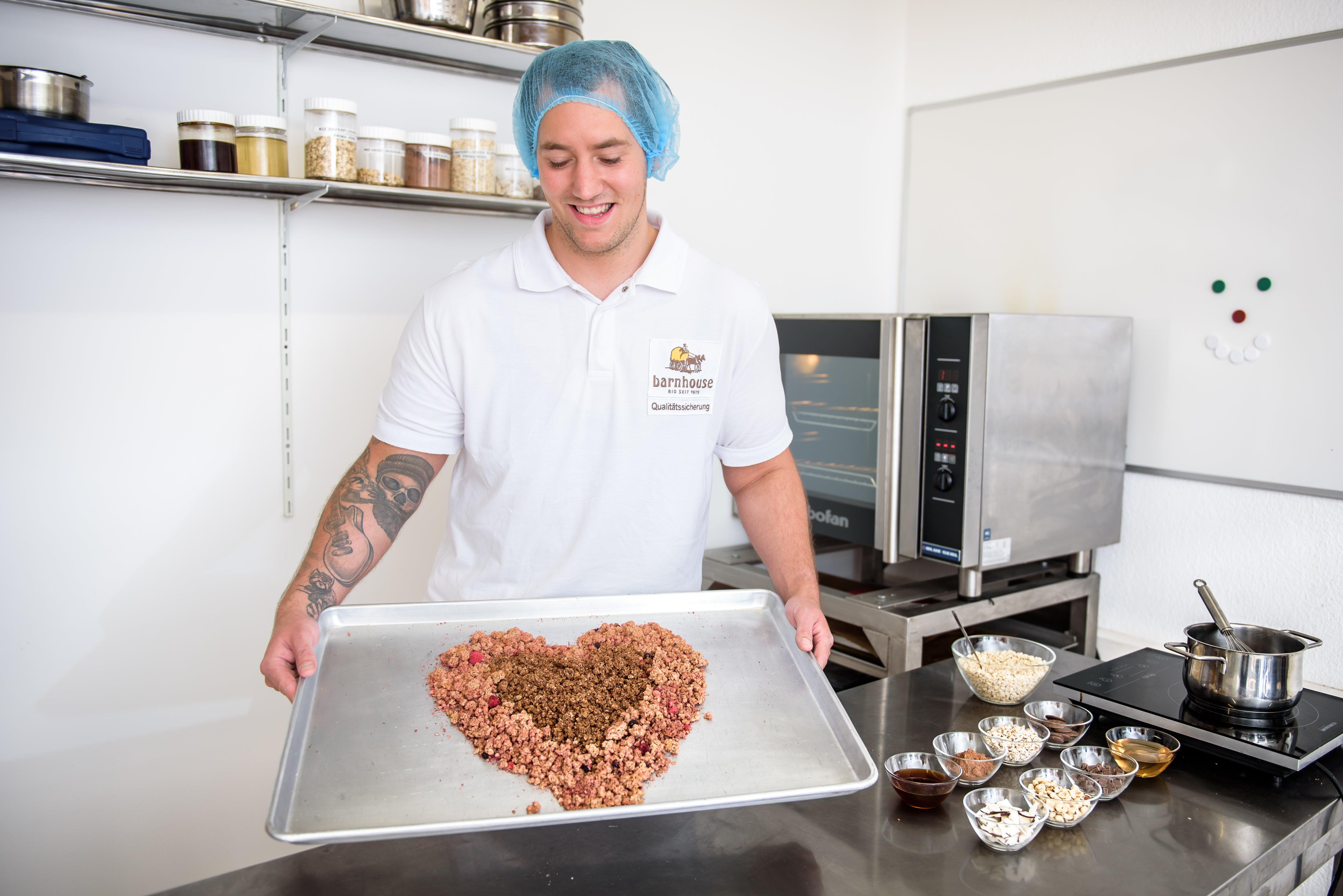 Produktentwickler Fabi hält in seiner Versuchsküche ein Blech mit einem Herz aus Krunchy, welches er glücklich anlacht. Außenrum stehen jede Menge Rohstoffe, die er für seine Produktentwicklung braucht. Im Hintergrund ein kleiner Ofen.