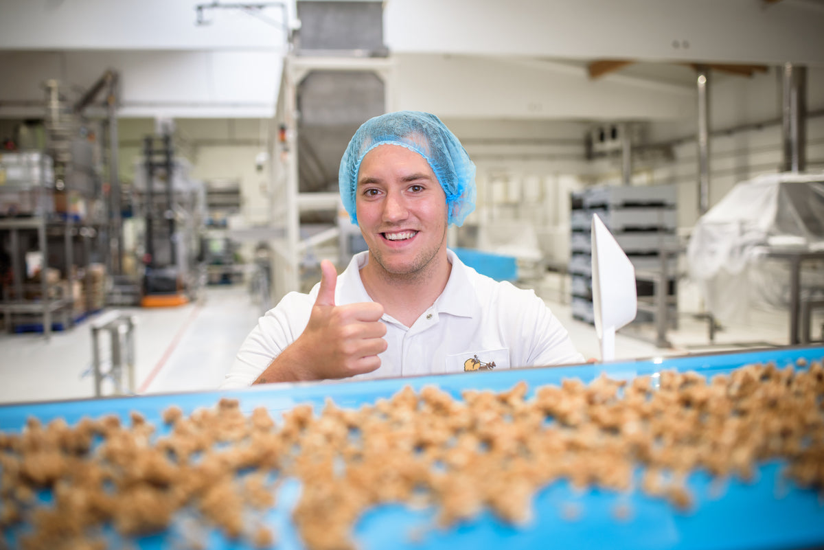 Produktentwickler Fabi der hinter einem Beförderungsband mit Krunchy steht und in die Kamera grinst. Mit der linken Hand hält er eine Handschaufel, während er mit der rechten Hand seinen Daumen nach oben streckt.