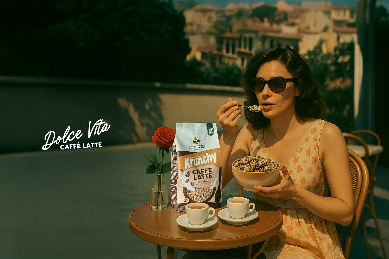 Dunkelhaarige Frau mit schwarzer Sonnenbrille und hellem Sommerkleid, die in einer Gasse mit italienischem Flair sitzt. An dem kleinen, runden Tisch vor ihr stehen zwei Tassen Espresso, eine durchsichtige Vase mit einer roten Rose und ein Beutel Krunchy Caffè Latte von Bio-Knuspermüsli Hersteller Barnhouse. In der linken Hand hält die Frau eine beige Schüssel mit diesem Müsli und mit der rechten Hand löffelt sie dieses. Im Hintergrund ist der Schriftzug "Dolce VIta - Caffè Latte" zu lesen.