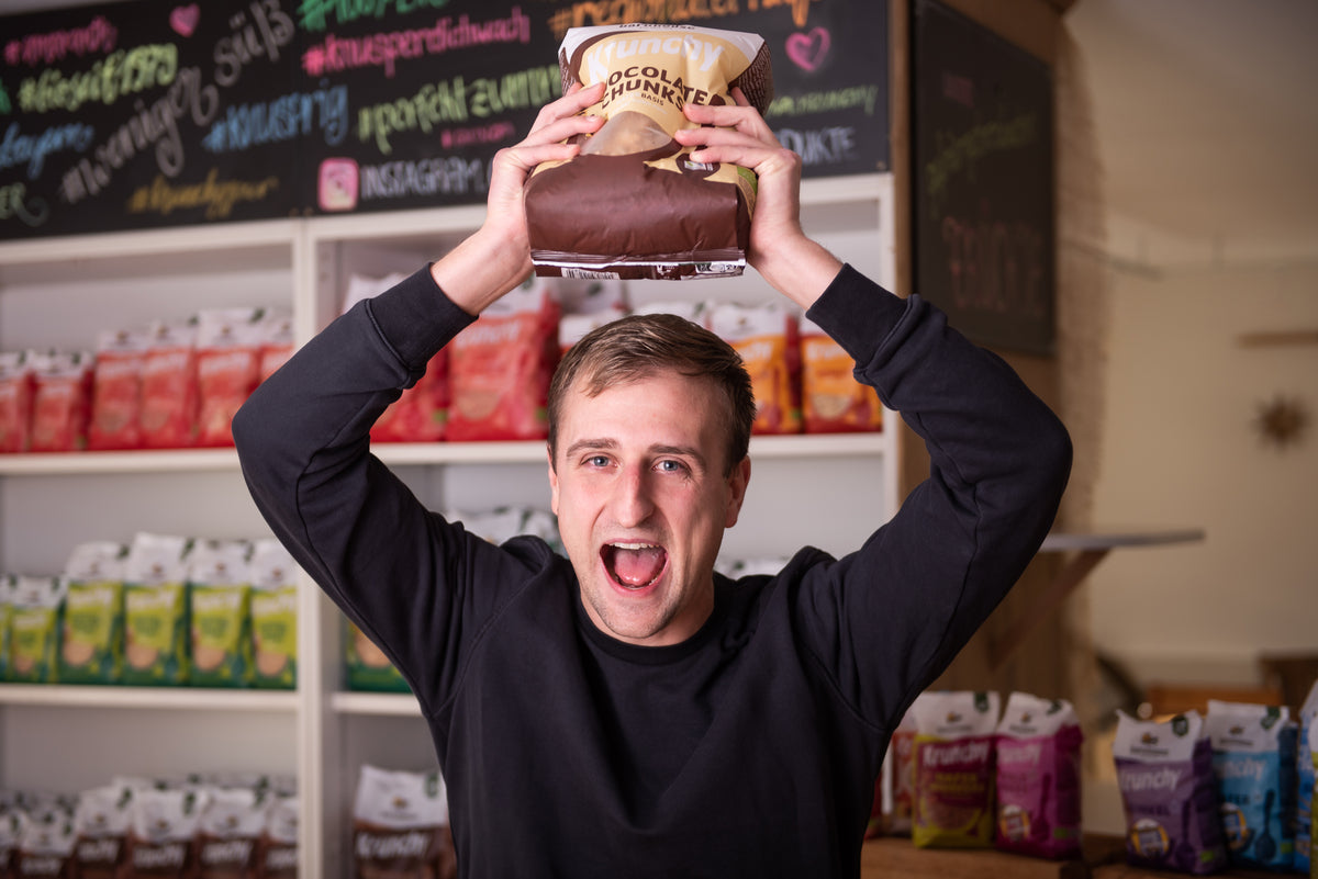 Spaßfoto: Christian - Leitung Qualität und Produktentwicklung - reißt jubelnd einen Beutel Krunchy Chocolate Chunks in die Höhe. Hinter ihm steht Regal mit Krunchy von Bio-Knuspermüsli-Hersteller Barnhouse.