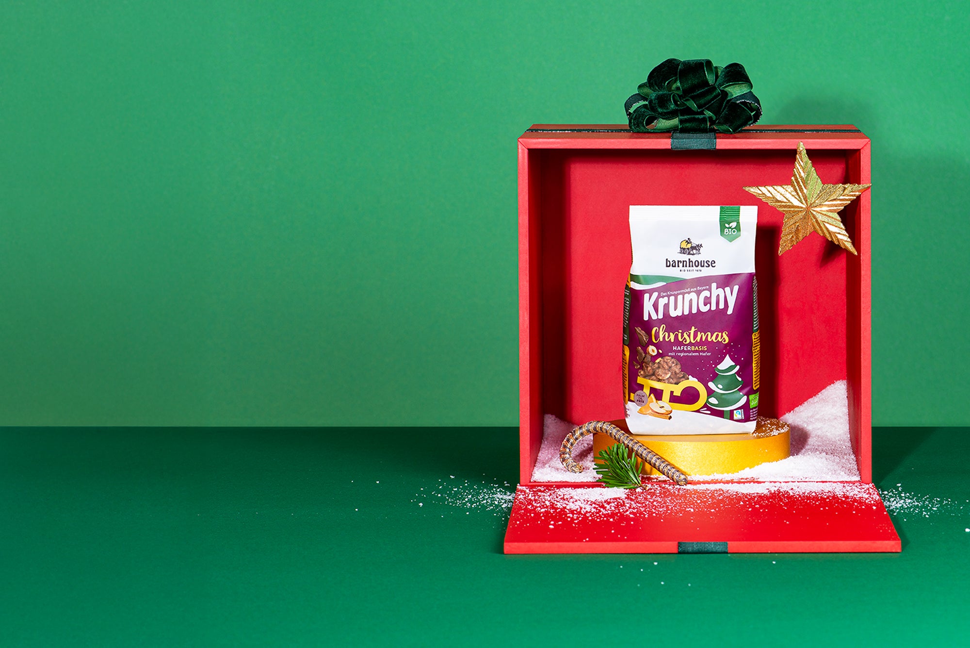 Beutel Krunchy Christmas auf goldenem Sockel mit goldenem Stern, goldener Zuckerstange und etwas Kunstschnee in einer roten Geschenkbox mit grüner Schleife vor grünem Hintergrund.