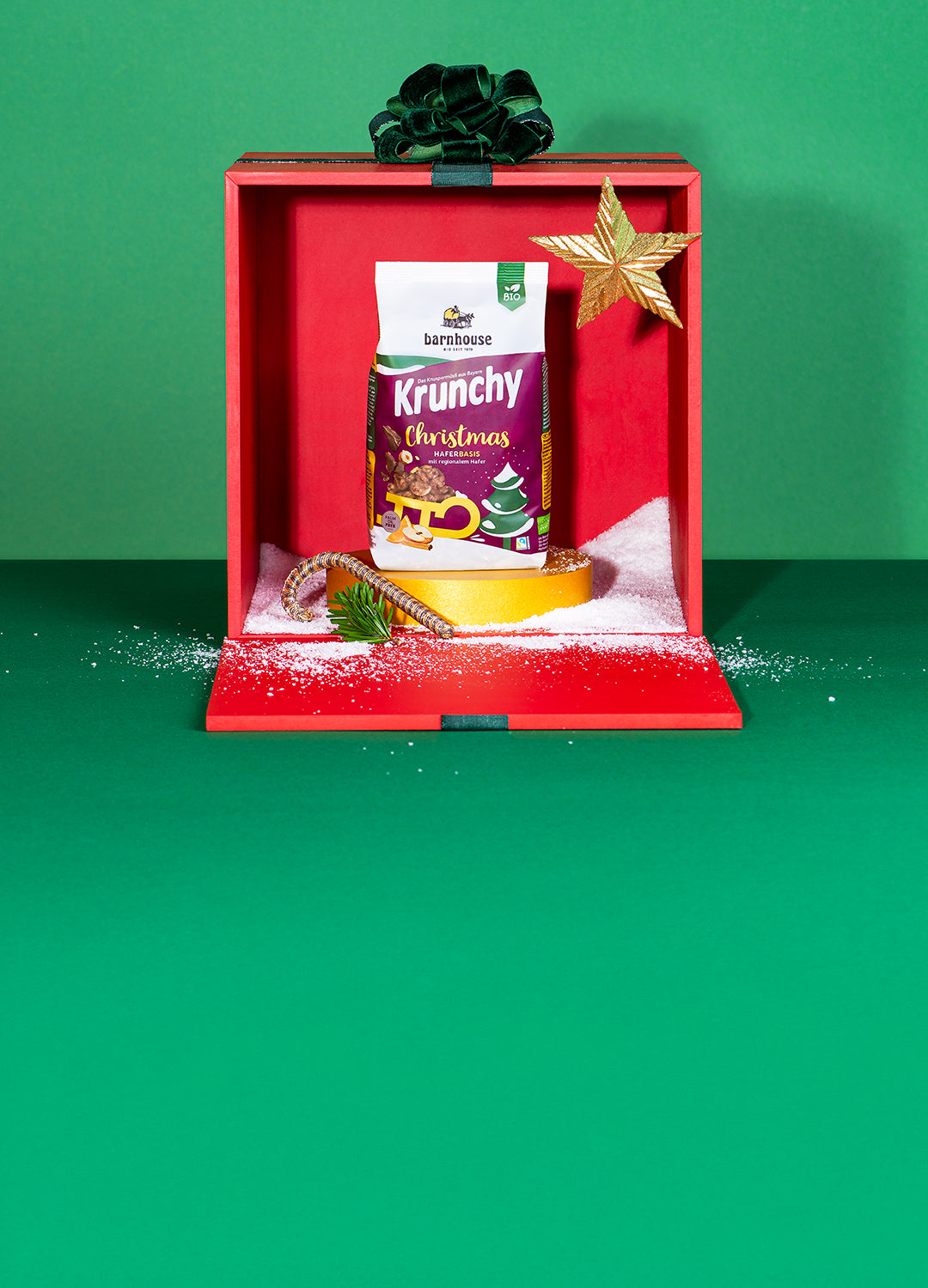Beutel Krunchy Christmas auf goldenem Sockel mit goldenem Stern, goldener Zuckerstange und etwas Kunstschnee in einer roten Geschenkbox mit grüner Schleife vor grünem Hintergrund.
