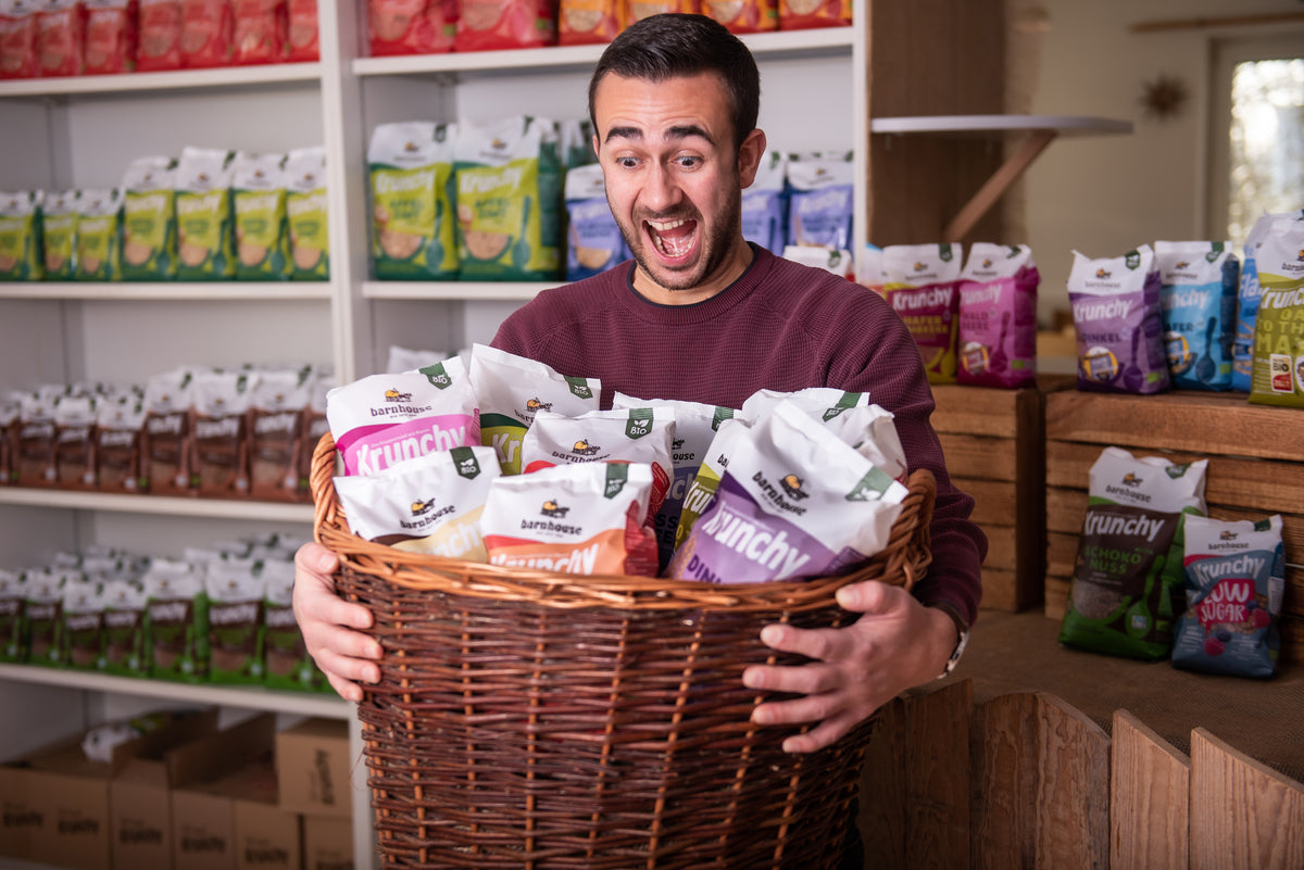 Spaßfoto: Daniel vom Onlinemarketing hält überglücklich einen geflochtenen Korb mit jeder Menge Krunchy. Hinter ihm steht eineRegal mit Krunchy von Bio-Knuspermüsli-Hersteller Barnhouse.