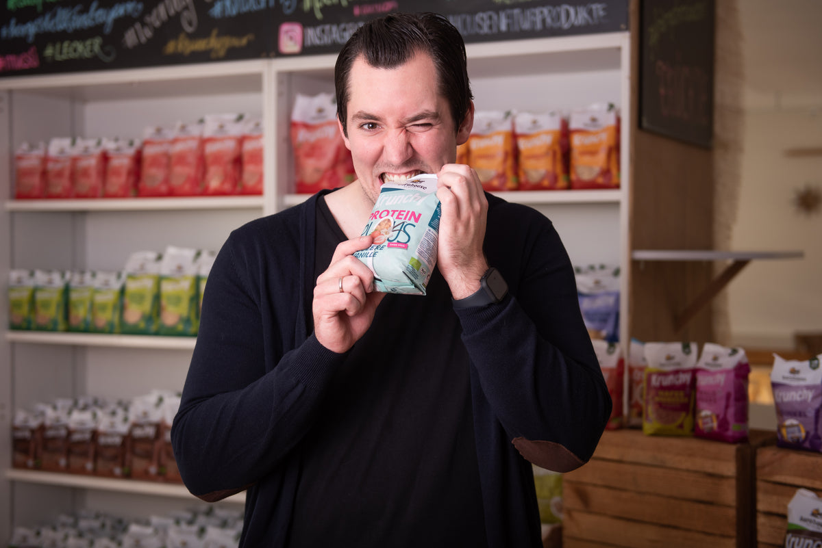 Spaßfoto: Produktentwickler Fabi beißt in einen geschlossen Beutel Krunchy Plus Protein. Hinter ihm steht ein Regal mit Krunchy von Bio-Knuspermüsli-Hersteller Barnhouse.