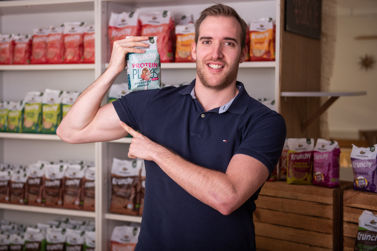 Spaßfoto: Fabi - Leiter Einkauf und Logistik - hält einen Beutel Krunchy Plus Protein auf seiner rechten Schulter und mit der linken Hand zeigt er auf seinen angespannten Bizeps. Hinter ihm steht ein Regal mit Krunchy von Bio-Knuspermüsli-Hersteller Barnhouse.