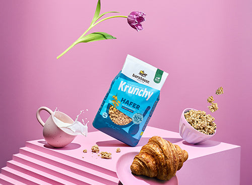 Blick voller Schwung von vorne auf ein rosa Treppe. Alle Elemente auf dem Bild "fliegen" durch die Luft: Blau-weißer Beutel Krunchy Hafer weniger Zucker von Barnhouse, fliederfarbene Schale mit diesem Bio-Knuspermüsli, Krunchy-Cluster, cremefarbene Tasse mit Milch, rosa Teller mit Croissant sowie eine Tulpe mit lila Blütenknospe.