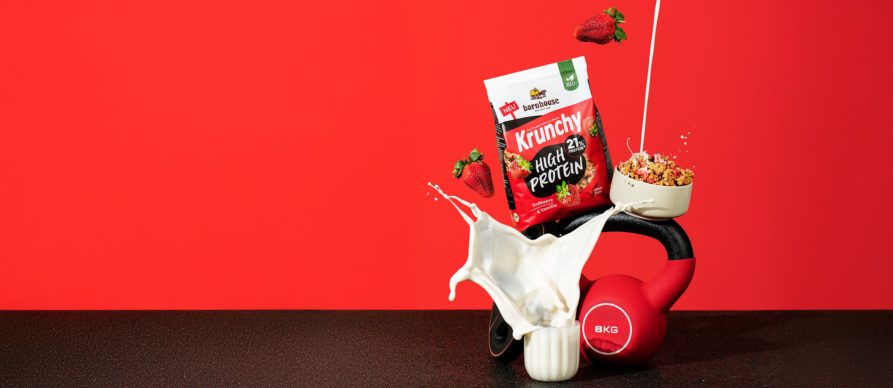 Blick von vorne auf einen Beutel Krunchy High Protein von Bio-Knuspermüsli-Hersteller Barnhouse, der auf einem Hantelgewicht und einer Kugelhantel steht. Davor steht ein Glas, aus dem Milch herausspritzt. Außerdem befindet sich noch eine Schüssel mit Krunchy High Protein auf der Kugelhantel, in die von oben ein Strahl Milch fließt. Zudem fliegen noch 2 Erdbeeren durch das Bild.