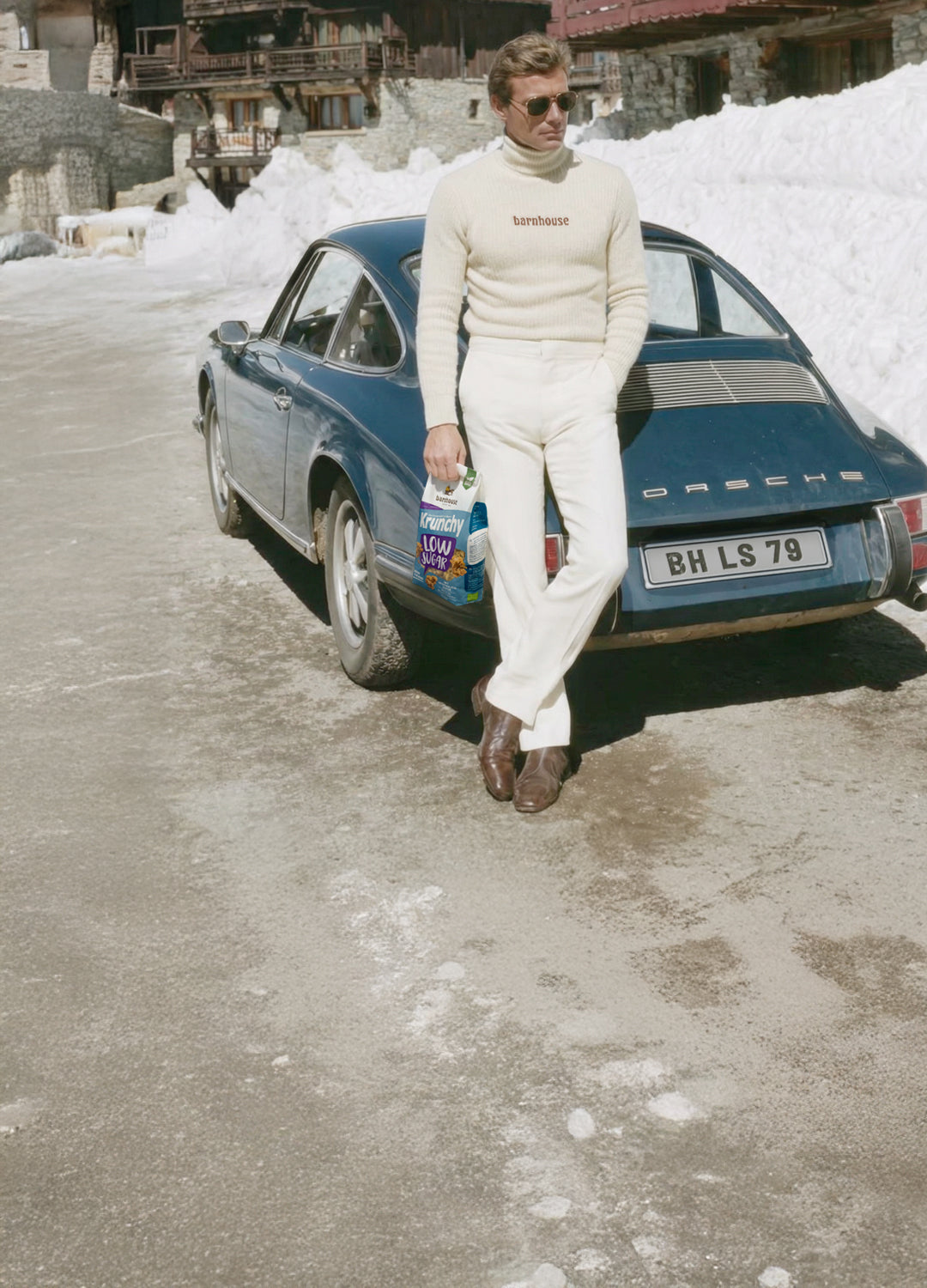 Mann mit weißer Hose, braunen Lederschuhen, Sonnenbrille und einem cremefarbenen Rollkragenpulli mit der Aufschrift "barnhouse" lehnt lässig an einem blauen Oldtimer-Porsche mit dem Kennzeichen "BH LS 79". In der rechten Hand hält er ein Krunchy Low Sugar Plain Grain. Im Hintergrund sind Almhütten und Schnee zu sehen.