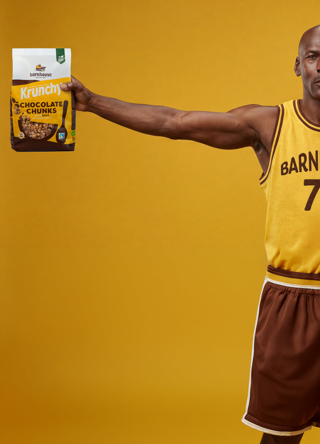 Basketballer mit gelbem Trikot mit der Aufschrift "Barnhouse 79". Seine Arme sind ausgestreckt. Mit der rechten Hand hält er einen Beutel Krunchy Chocolate Chunks, mit der linken eine Krunchy-Bowl mit Schokolade und Himbeeren.