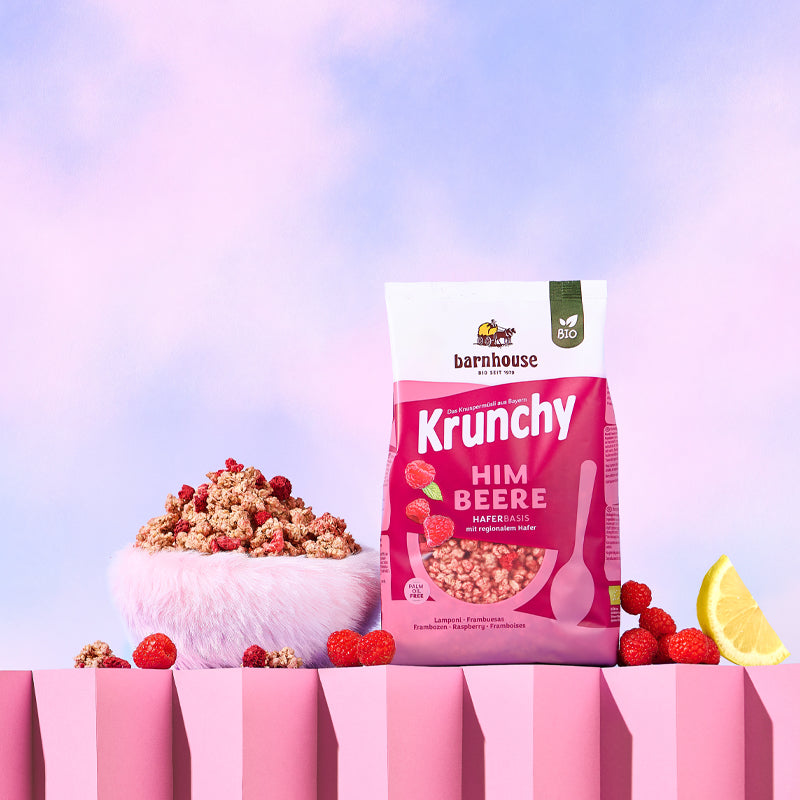 Blick von vorne auf den beerenfarbigen, rosa-weißer Beutel Krunchy Himbeere von Bio-Knuspermüsli-Hersteller Barnhouse, der auf einem gezackten, rosafarbenen Sockel steht. Links daneben steht eine Schüssel mit diesem Müsli, die von Himbeeren und Krunchy-Clustern umgeben ist. Rechts daneben liegen ein paar Himbeeren sowie ein Achtel einer Zitrone. Der Hintergrund ist hell.