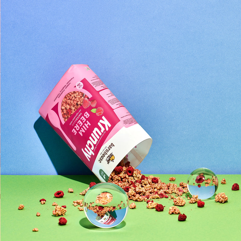 Blick von vorne auf den beerenfarbigen, rosa-weißen Beutel Krunchy Himbeere von Bio-Knuspermüsli-Hersteller Barnhouse. Der Beutel ist auf den Kopf gedreht und geöffnet, wodurch jede Menge Krunchy-Cluster und Himbeeren herausrieseln und auf den grünen Untergrund fallen. Zwischendrin liegen 2 Glaskugeln, die Elemente des Bildausschnittes spiegeln. Die Wand im Hintergrund ist blau.