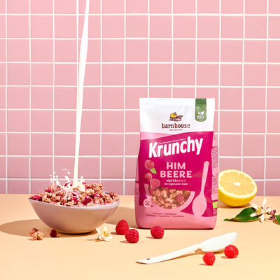 Blick von vorne auf den beerenfarbigen, rosa-weißer Beutel Krunchy Himbeere von Bio-Knuspermüsli-Hersteller Barnhouse, der auf einem beigen Tisch steht. Links daneben steht eine Schüssel mit diesem Müsli, in die von oben ein Milchstrahl fließt. Daneben liegen eine halbe Zitrone, Himbeeren, Krunchy-Cluster sowie ein weißer Löffel und zwei weiße Blüten. Im Hintergrund ist eine rosa geflieste Wand zu sehen.