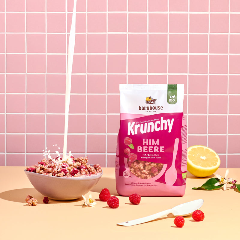 Blick von vorne auf den beerenfarbigen, rosa-weißer Beutel Krunchy Himbeere von Bio-Knuspermüsli-Hersteller Barnhouse, der auf einem beigen Tisch steht. Links daneben steht eine Schüssel mit diesem Müsli, in die von oben ein Milchstrahl fließt. Daneben liegen eine halbe Zitrone, Himbeeren, Krunchy-Cluster sowie ein weißer Löffel und zwei weiße Blüten. Im Hintergrund ist eine rosa geflieste Wand zu sehen.