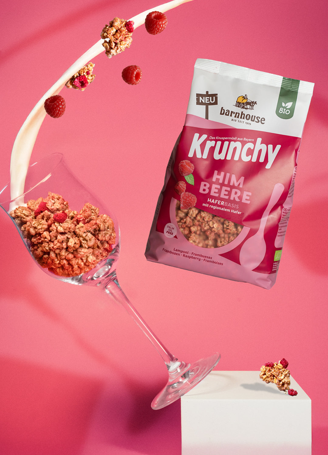 Beutel Krunchy Himbeere, der rechts neben einem Weinglas schwebt, das links von einem weißen Sockel fällt. Das Weinglas ist mit Clustern des Himbeer-Müslis befüllt. Vom oberen Bildrand fließt ein Milchstrahl ins Glas und weitere Cluster sowie Himbeeren fliegen herum.
