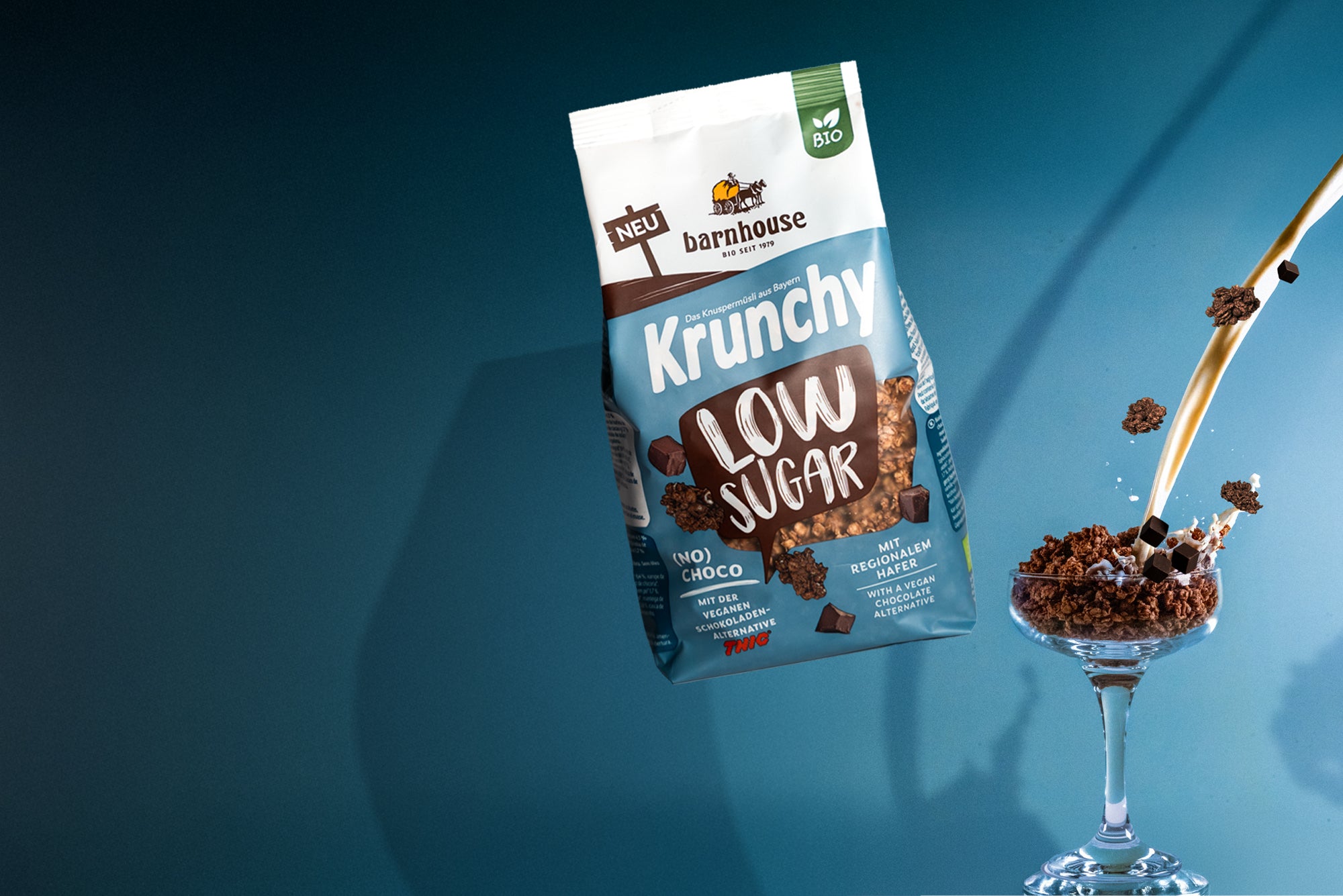 Beutel Krunchy Low Sugar (No) Choco, der über einem Glas schwebt, das mit Knuspermüsli-Clustern befüllt ist. Vom rechten Bildrand fließt ein Milchstrahl ins Glas und "Schoko"-Cluster fliegen herum.