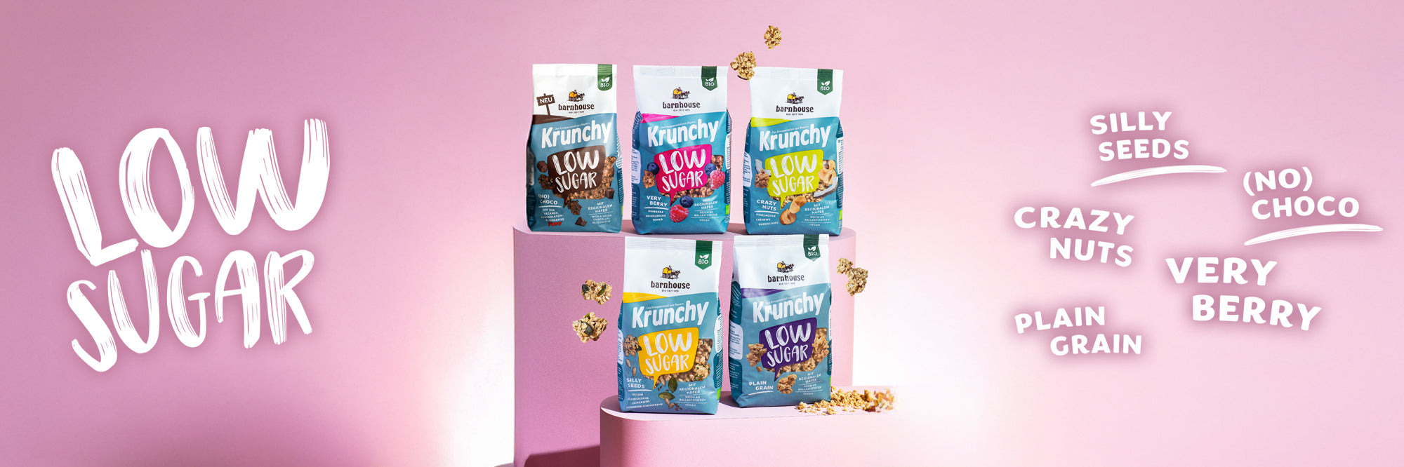 Blick von vorne auf die fünf Low Sugar Krunchys Plain Grain, Crazy Nuts, Very Berry (No) Choco und Silly Seeds, die auf zwei verschiedenen rosa Sockeln stehen. Außenrum fliegen Cluster. Links steht Low Sugar, rechts die fünf Sortennamen.