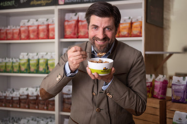 Spaßfoto: Geschäftsführer Martin Eras hält in der linken Hand eine Barnhouse-Schüssel mit Krunchy und mit der rechten Hand führt er den befüllten Löffel lächelnd zu seinem Mund. Hinter ihm steht ein Regal mit Krunchy von Bio-Knuspermüsli-Hersteller Barnhouse.