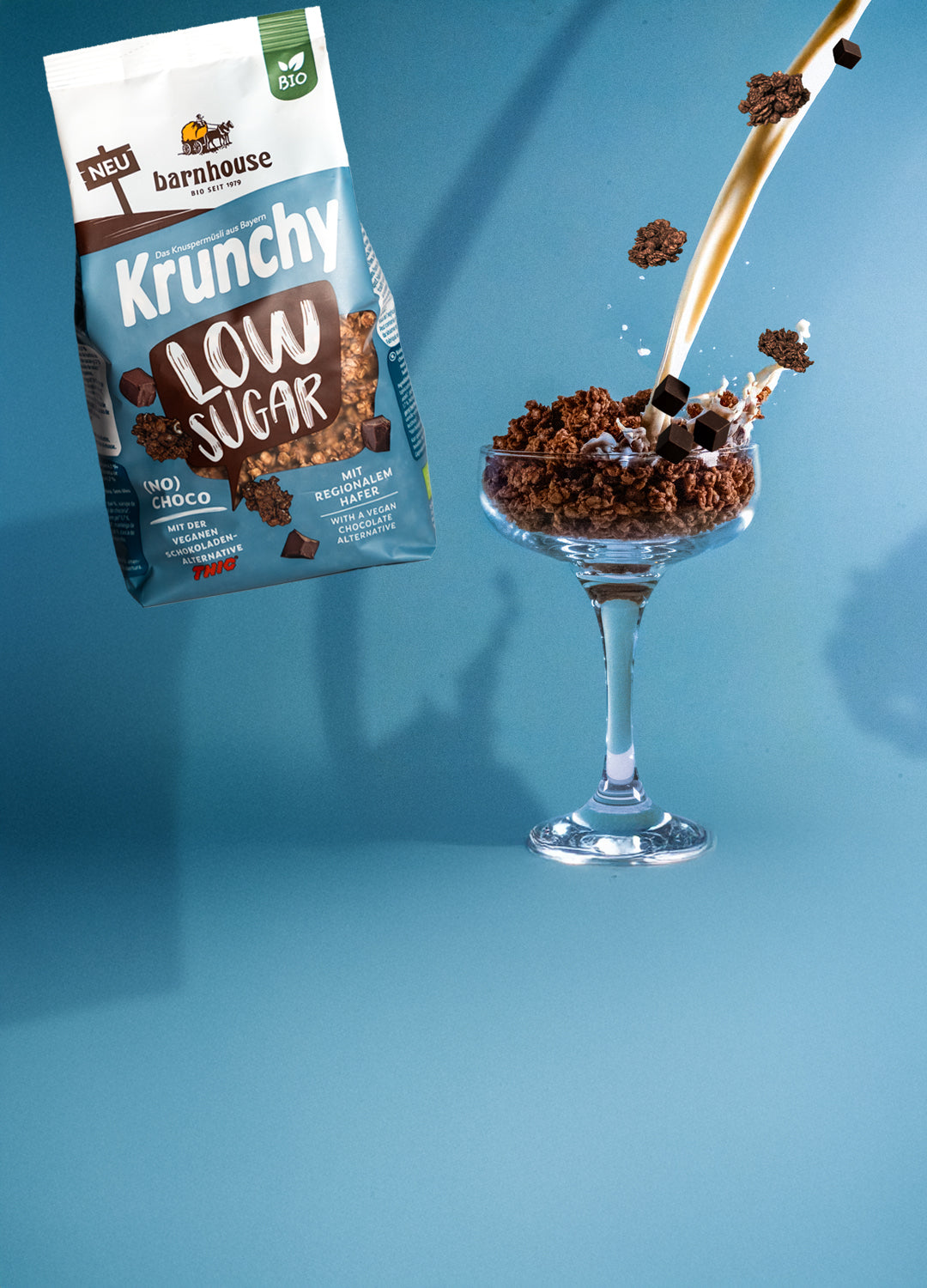 Beutel Krunchy Low Sugar (No) Choco, der über einem Glas schwebt, das mit Knuspermüsli-Clustern befüllt ist. Vom rechten, oberen Bildrand fließt ein Milchstrahl ins Glas und "Schoko"-Cluster fliegen herum.